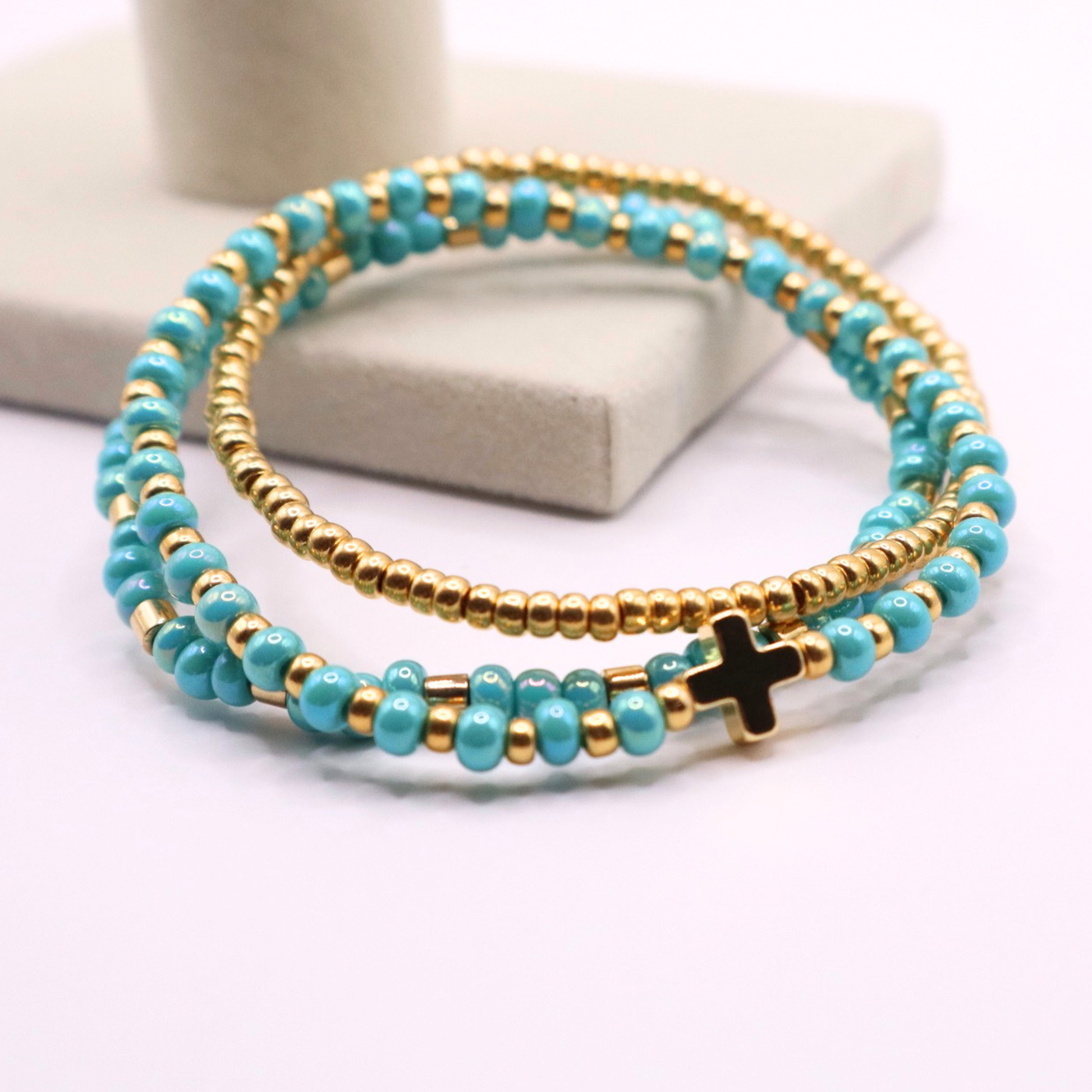 Turquoise and gold beaded bracelet stack with a cross charm on a white surface