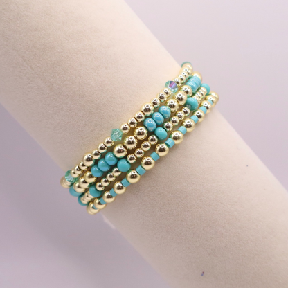 Layered bracelet stack with turquoise Preciosa seed beads, shimmering crystal accents, and gold-tone hematite beads on a clean white background.