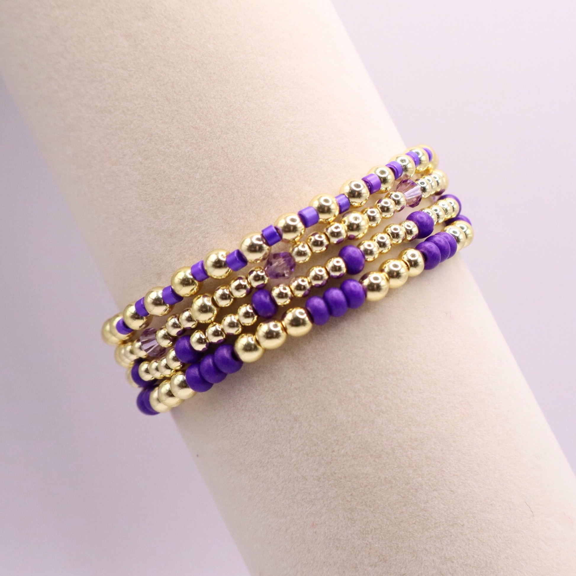 Elegant stack of stretch bracelets featuring rich purple Preciosa seed beads, faceted crystal details, and gold-tone hematite accent beads on a clean white background.