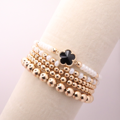 Set of 5 of gold toned beaded bracelets with white seed beads and hematite gold accents. One bracelet features a black clover charm. The bracelets are displayed on a white background.