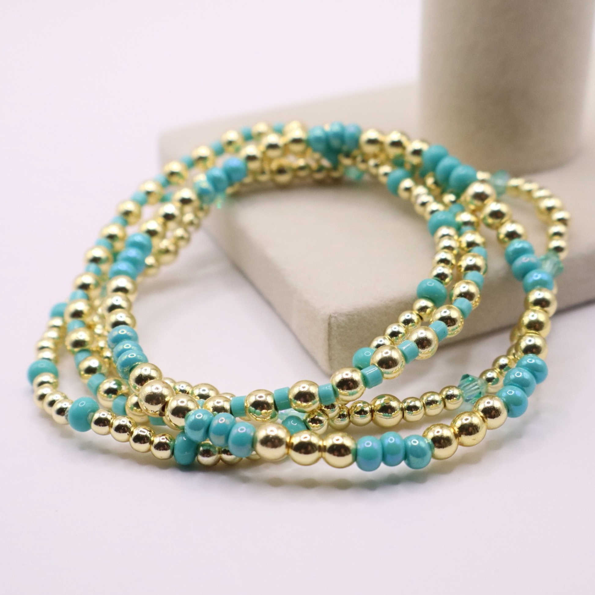 Layered bracelet stack with turquoise Preciosa seed beads, shimmering crystal accents, and gold-tone hematite beads on a clean white background.