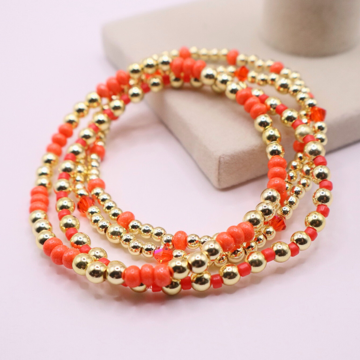 Stack of handmade stretch bracelets featuring vibrant coral Preciosa seed beads, faceted crystal accents, and polished gold-tone hematite beads on a clean white background.