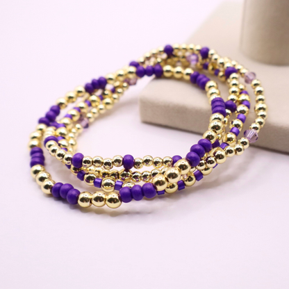 Elegant stack of stretch bracelets featuring rich purple Preciosa seed beads, faceted crystal details, and gold-tone hematite accent beads on a clean white background.