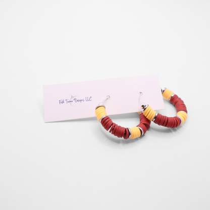 Burgundy and gold heishi bead hoop earrings with gold accents hanging on a minimalist jewelry stand against a white background