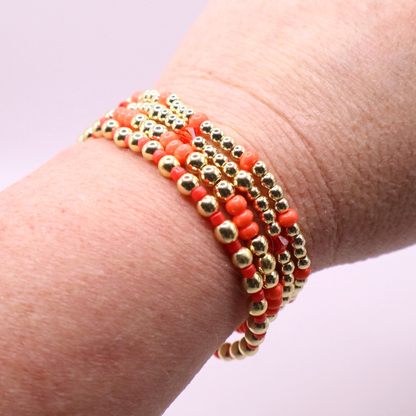 Stack of handmade stretch bracelets featuring vibrant coral Preciosa seed beads, faceted crystal accents, and polished gold-tone hematite beads on a clean white background.