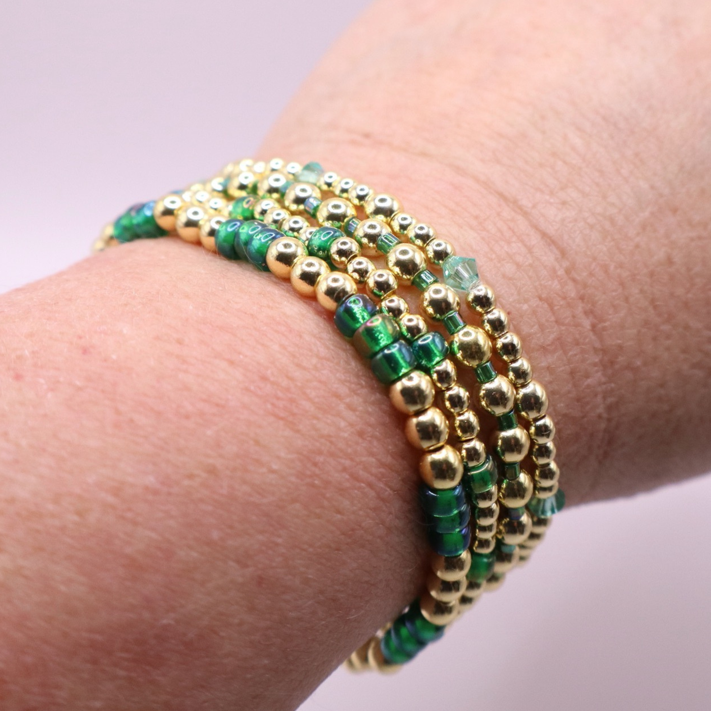 Handmade bracelet stack with green Preciosa seed beads, iridescent crystal accents, and polished gold-tone hematite beads on a clean white background.