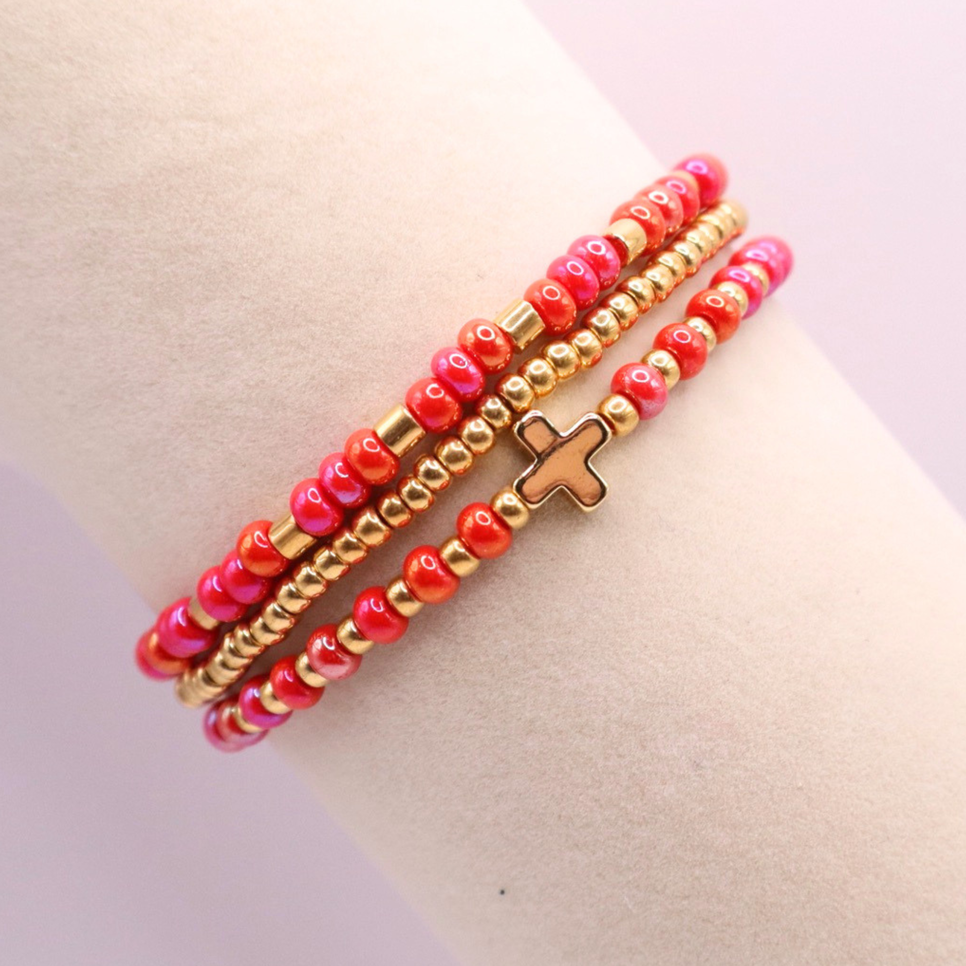 Pink beaded bracelet stack with gold accents and a cross charm on a light background
