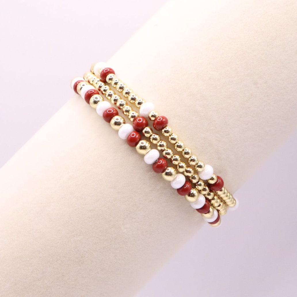 Three crimson, white and gold beaded bracelet on a wrist against a light background