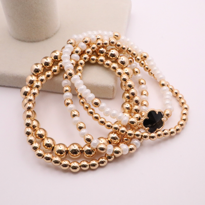Set of 5 of gold toned beaded bracelets with white seed beads and hematite gold accents. One bracelet features a black clover charm. The bracelets are displayed on a white background.