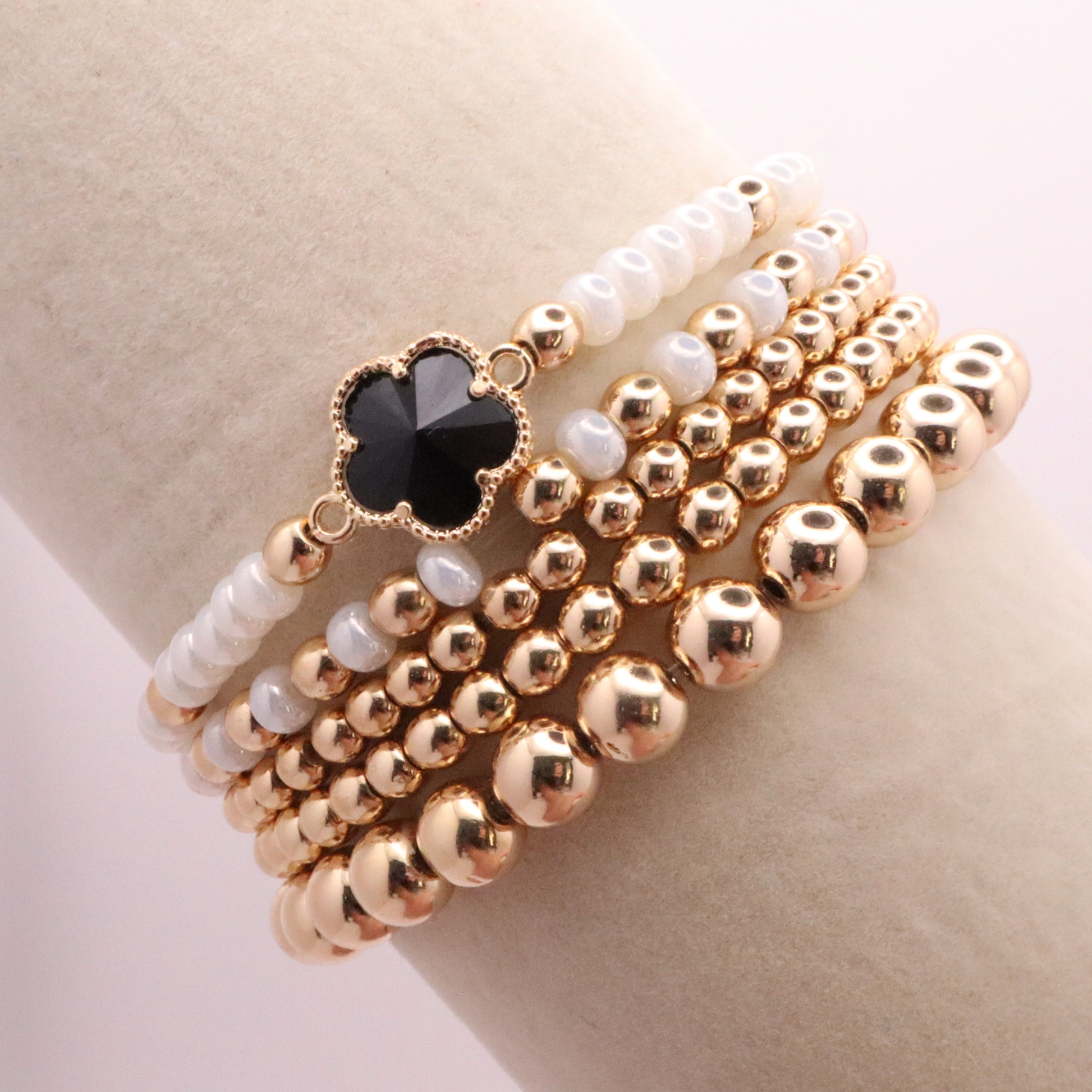 Set of 5 of gold toned beaded bracelets with white seed beads and hematite gold accents. One bracelet features a black clover charm. The bracelets are displayed on a white background.