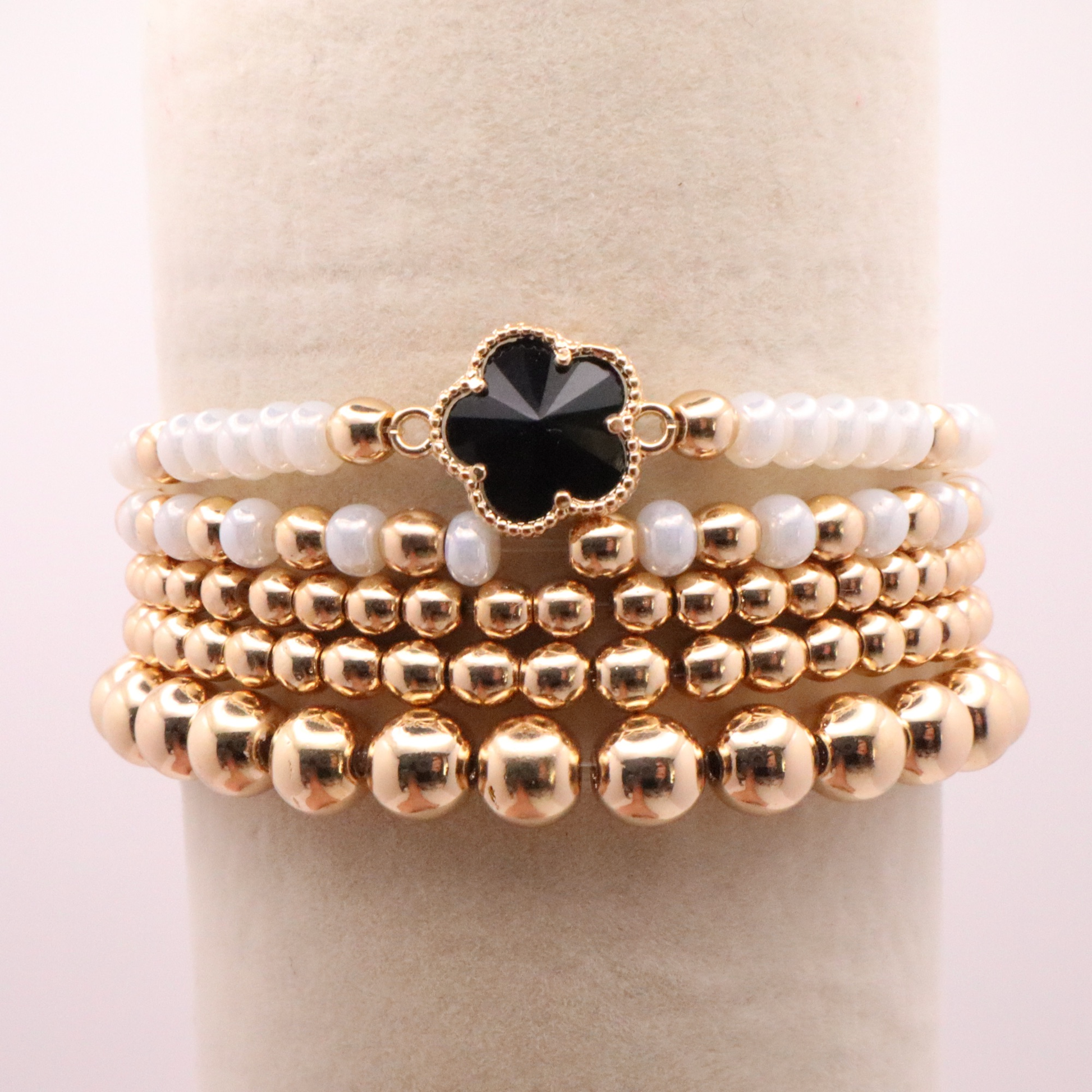 Set of 5 of gold toned beaded bracelets with white seed beads and hematite gold accents. One bracelet features a black clover charm. The bracelets are displayed on a white background.