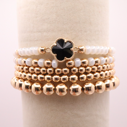 Set of 5 of gold toned beaded bracelets with white seed beads and hematite gold accents. One bracelet features a black clover charm. The bracelets are displayed on a white background.
