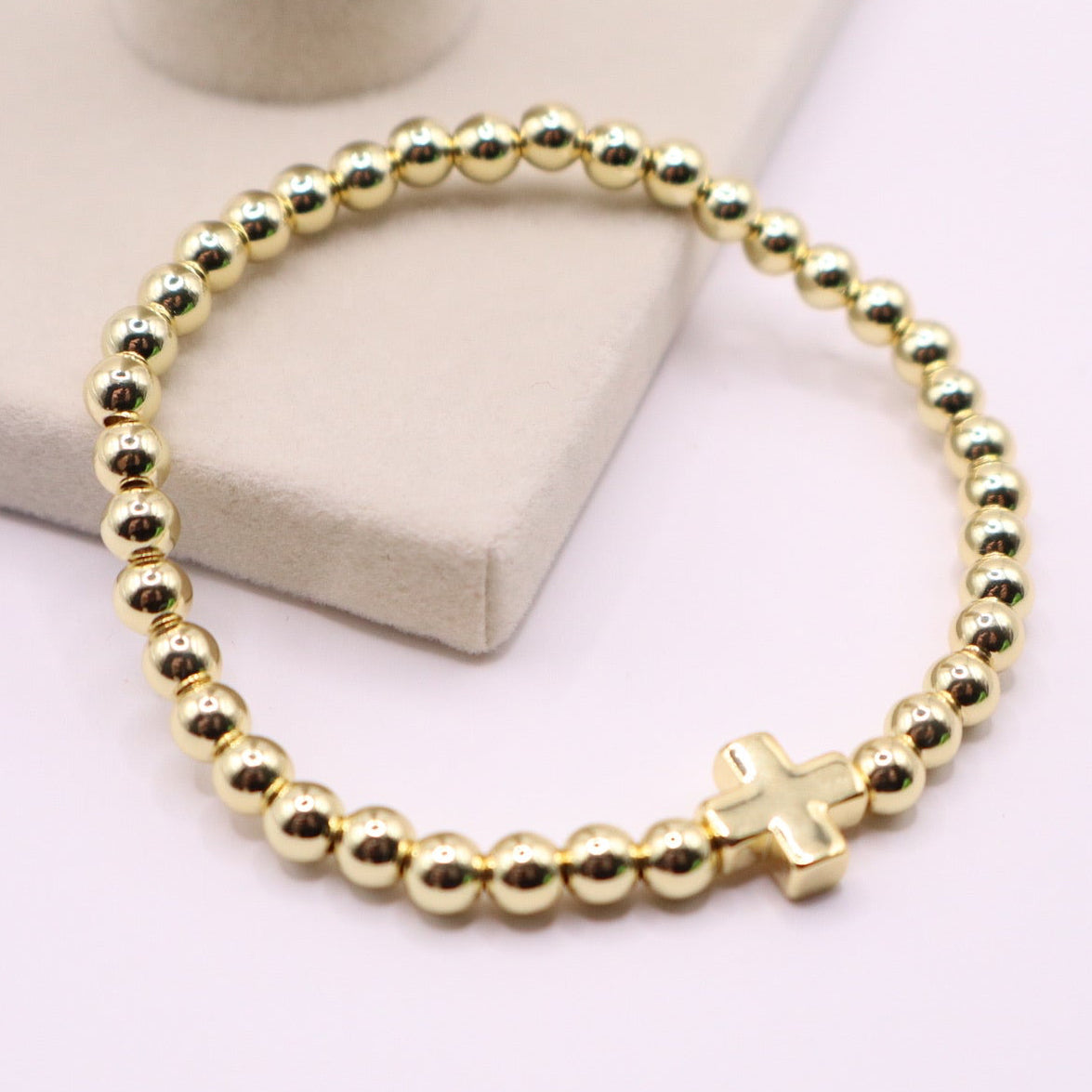 18K gold filled beaded stretch bracelet with a small polished cross charm, displayed on a soft neutral jewelry stand with a clean, minimal background.
