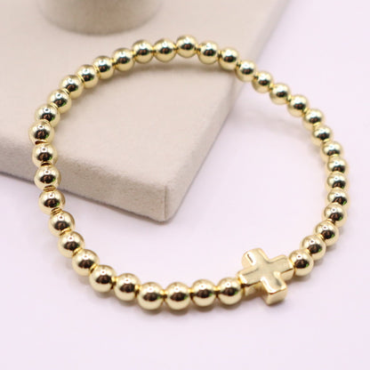 18K gold filled beaded stretch bracelet with a small polished cross charm, displayed on a soft neutral jewelry stand with a clean, minimal background.