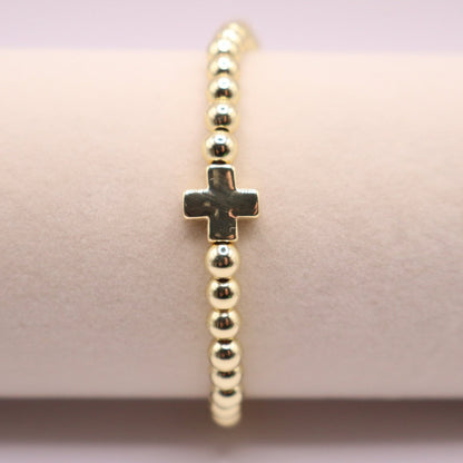 Centered view of a 18k gold filled beaded bracelet with polished round gold beads and a small cross charm, displayed on a blush-toned bracelet stand against a soft pastel background.
