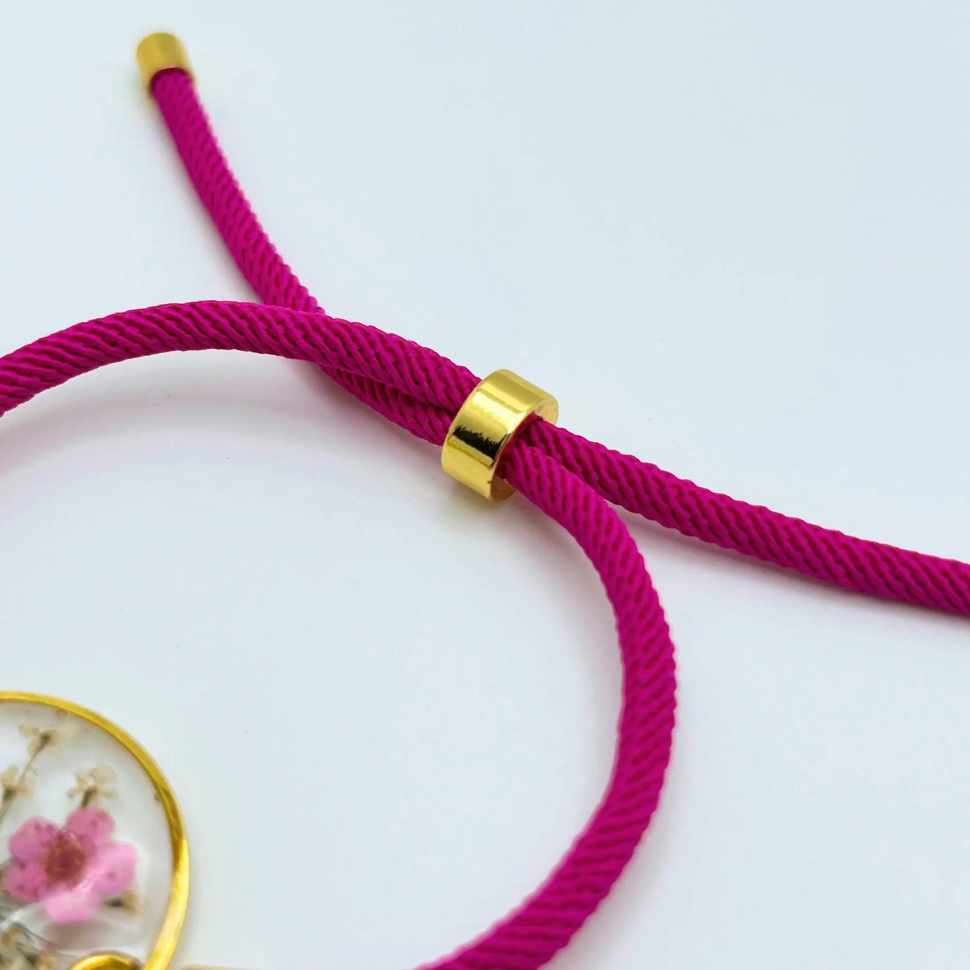 Close-up of a magenta-colored bracelet cord with gold-colored metal accents. The bracelet features a sliding knot for adjustable sizing. The cord is thick and appears to be made of a woven material.