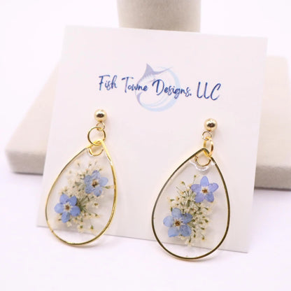 Pair of clear resin earrings with blue flowers and delicate white florals framed in gold teardrop shape shown on display card with Fish Towne Designs branding