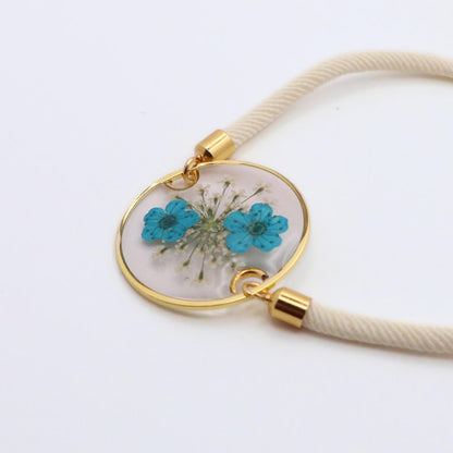 Handmade blue flower resin bracelet with gold round bezel and white adjustable cord on white background
