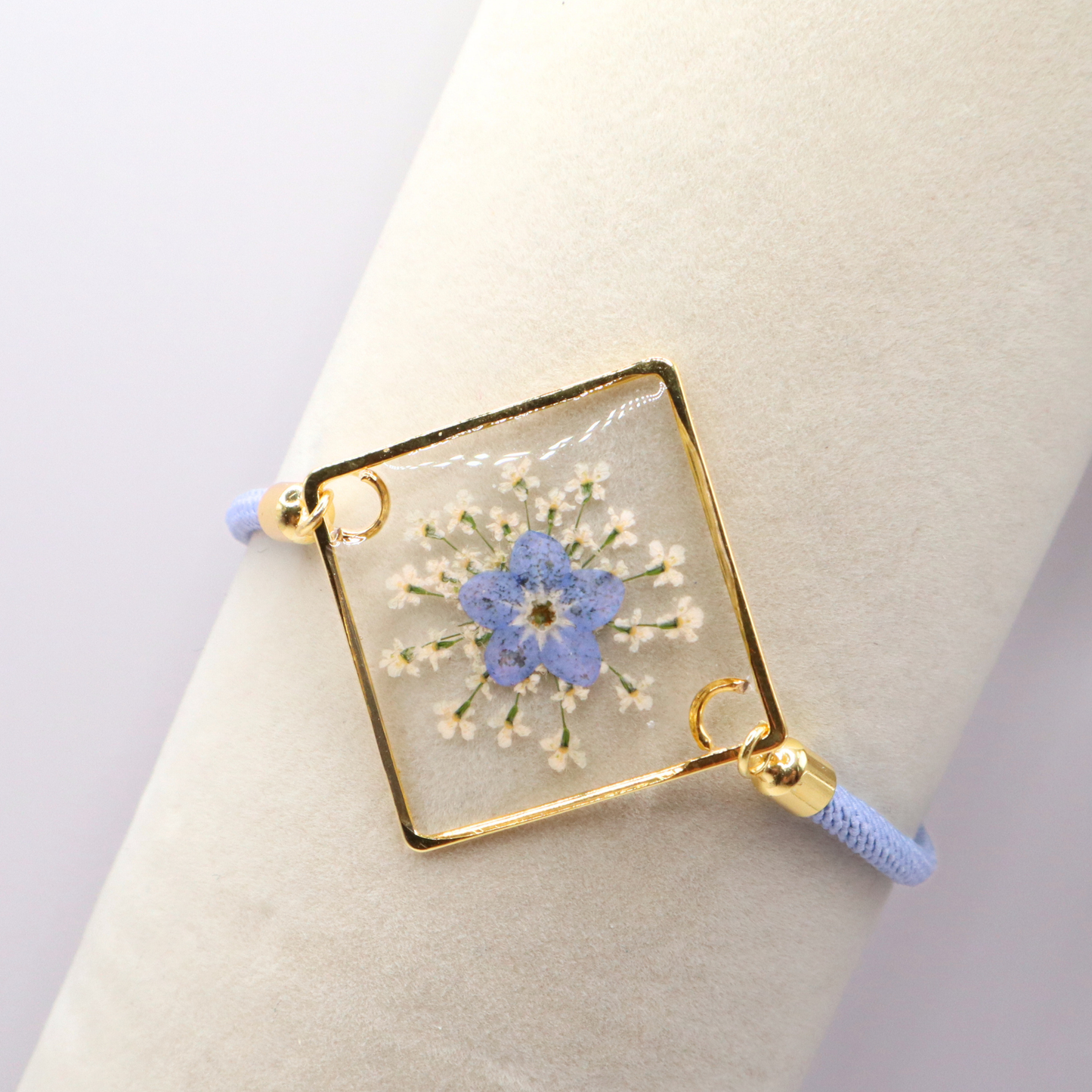 Dainty blue forget-me-not flower resin bracelet with gold frame and adjustable cord displayed on a display rack.  It is available with gold or silver tone accents.

