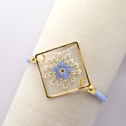 Dainty blue forget-me-not flower resin bracelet with gold frame and adjustable cord displayed on a display rack.  It is available with gold or silver tone accents.

