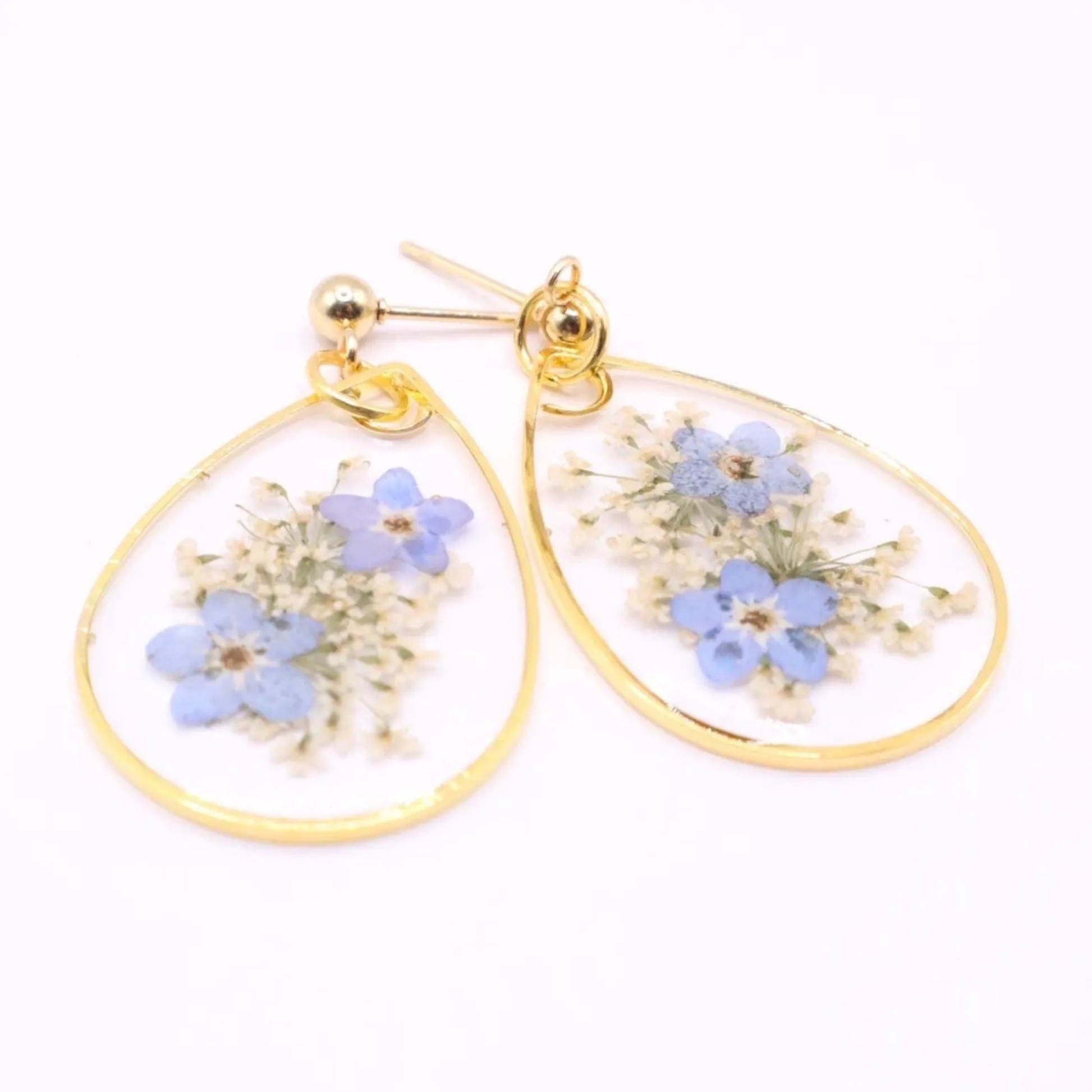 Detailed view of resin earrings with real blue flowers and white filler blooms in clear teardrop design with gold trim