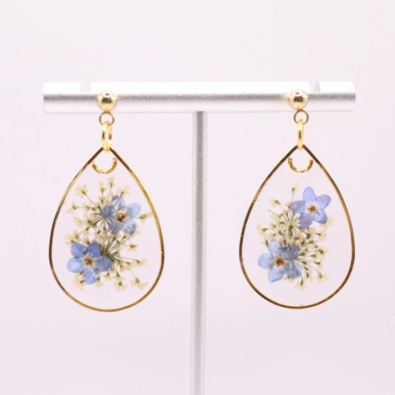 Gold framed resin earrings with blue dried flowers and white accents displayed on stand with soft neutral background