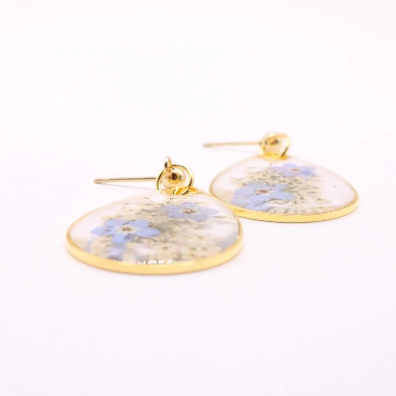 Flat lay of handmade resin earrings with blue pressed flowers in clear teardrop shape with gold edge and stud posts