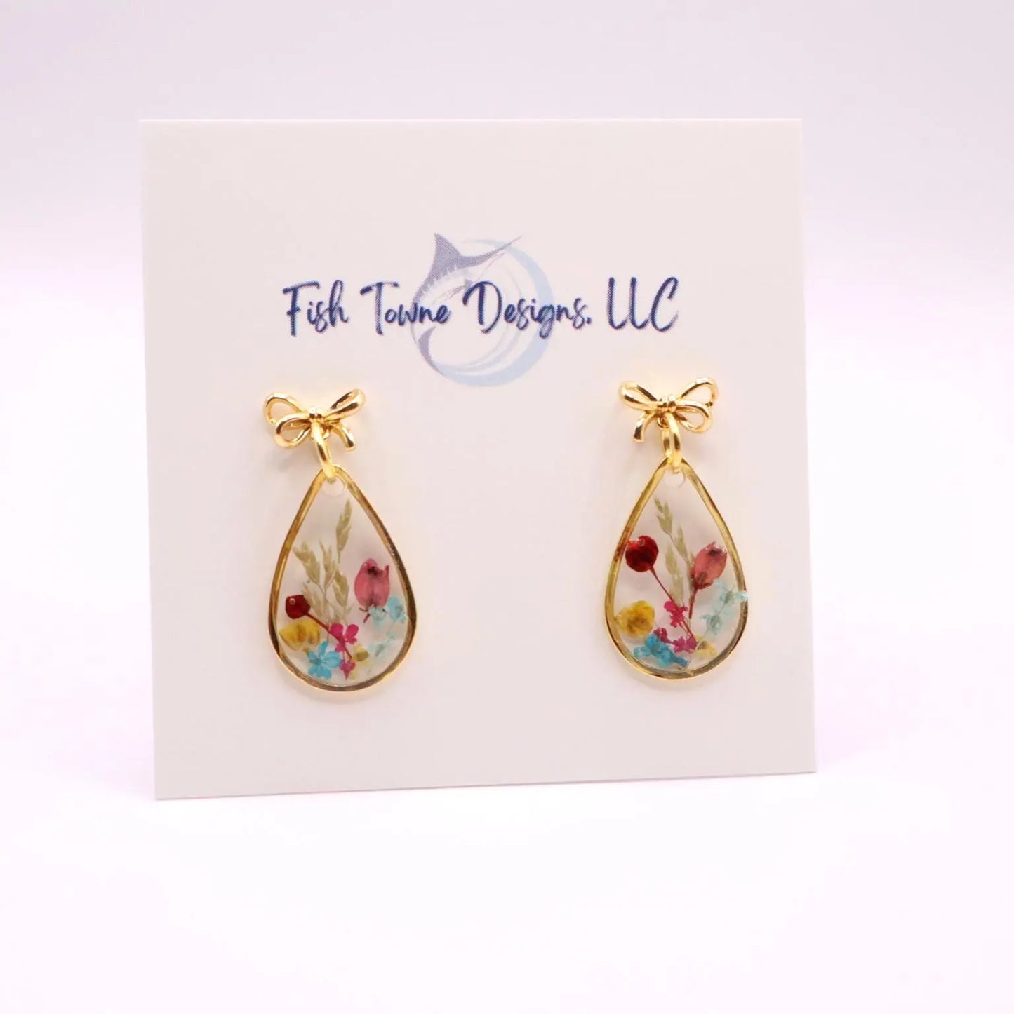 Gold-toned teardrop earrings with an 18k gold plated bow detail at the top. Each earring features a clear resin interior filled with colorful dried flowers. The earrings are displayed on a white card with the text "Fish Towne Designs, LLC".