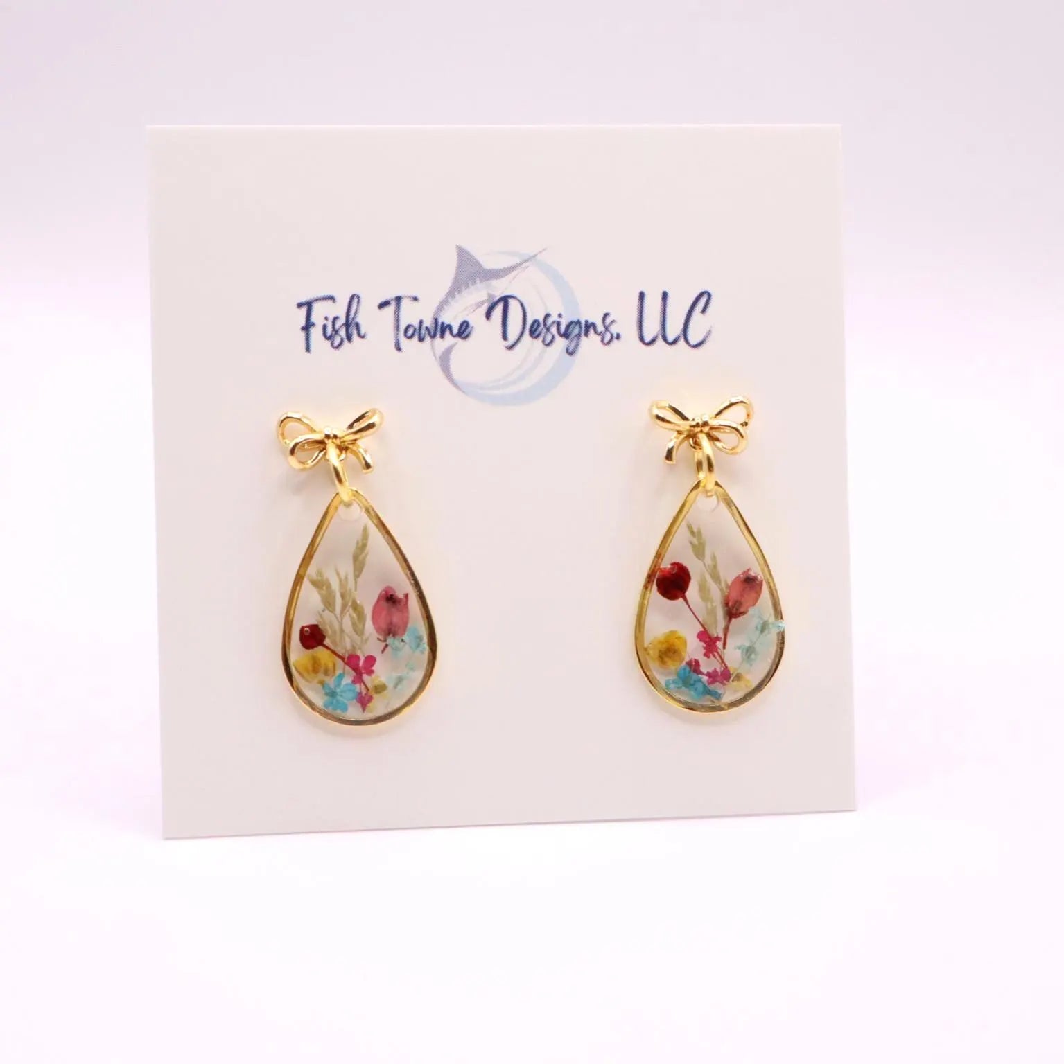 Gold-toned teardrop earrings with an 18k gold plated bow detail at the top. Each earring features a clear resin interior filled with colorful dried flowers. The earrings are displayed on a white card with the text "Fish Towne Designs, LLC".