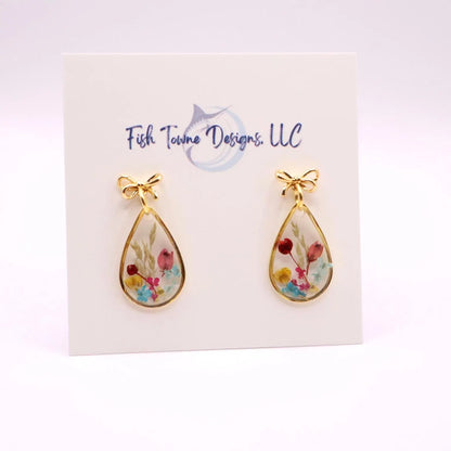 Gold-toned teardrop earrings with an 18k gold plated bow detail at the top. Each earring features a clear resin interior filled with colorful dried flowers. The earrings are displayed on a white card with the text "Fish Towne Designs, LLC".