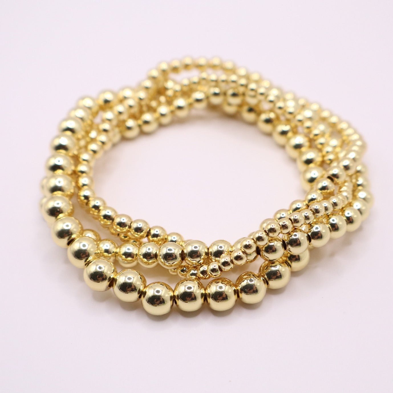 Four-piece 18K gold filled bracelet stack featuring polished round beads in varying sizes, arranged in a layered circle on a soft neutral background.