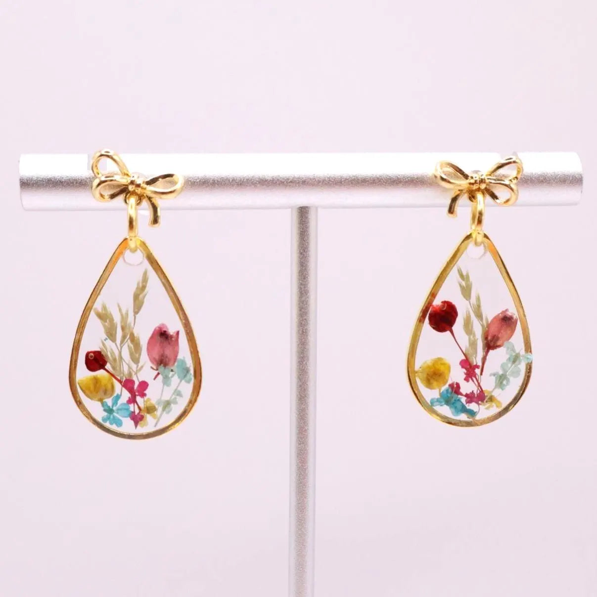 A pair of gold-toned teardrop earrings. Each earring features a clear resin teardrop shape with dried flowers in shades of red, pink, yellow, and blue. A small 18k gold plated bow tops each earring.