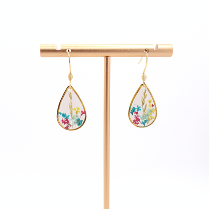 Handmade teardrop floral resin earrings with gold frames hanging on a gold stand against a white background