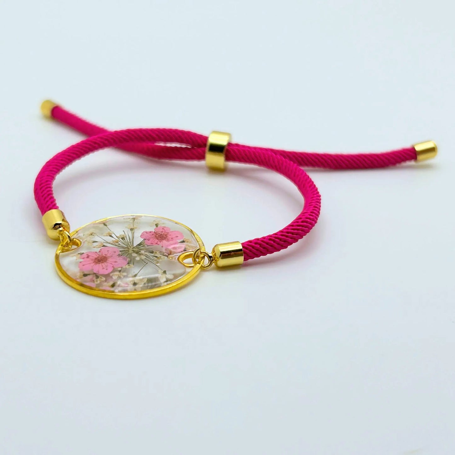 A hot pink adjustable bracelet with a gold-colored circular pendant. The pendant is filled with dried pink flowers and clear resin. The bracelet's cord is a vibrant pink, and the adjustable clasp is gold-colored.