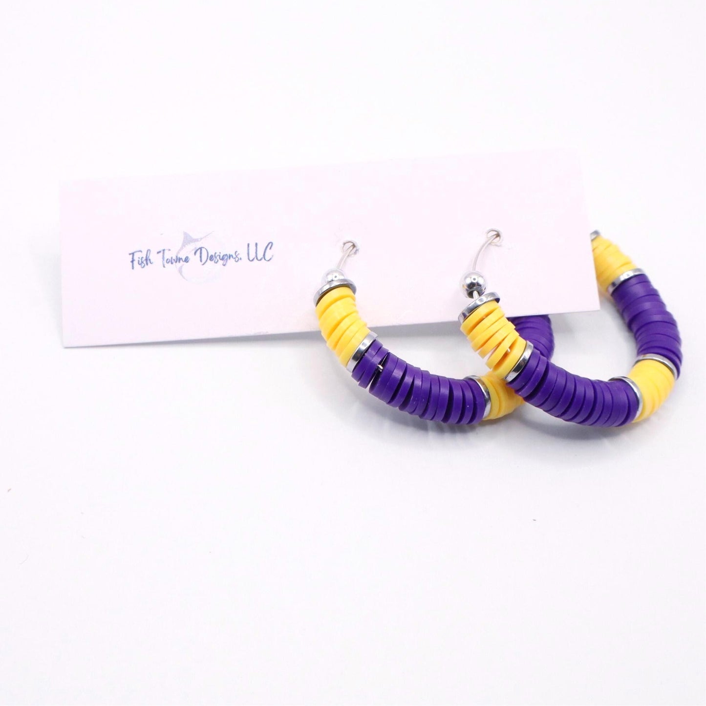 Purple and gold heishi bead hoop earrings with gold accents hanging on a minimalist jewelry stand against a white background