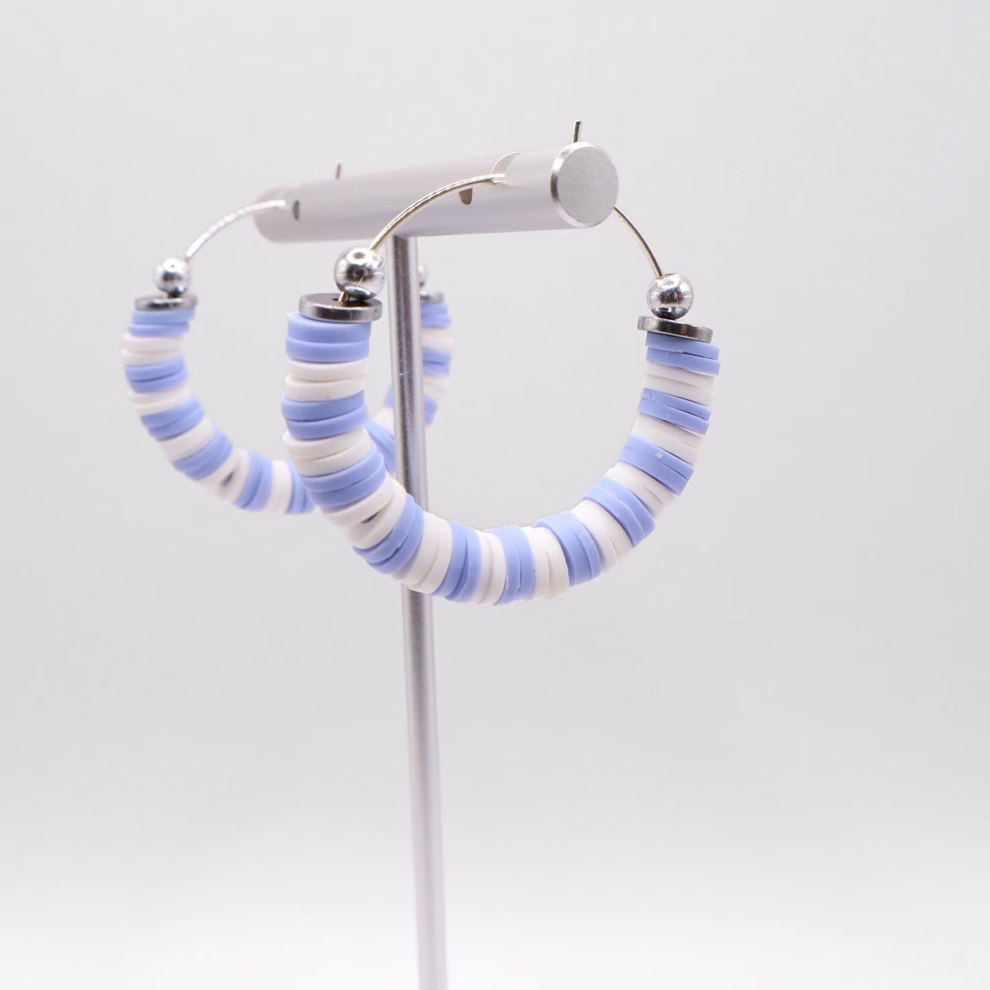 Close-up of light blue and white heishi bead hoop earrings with silver accents on a jewelry stand, highlighting bead detail and pattern