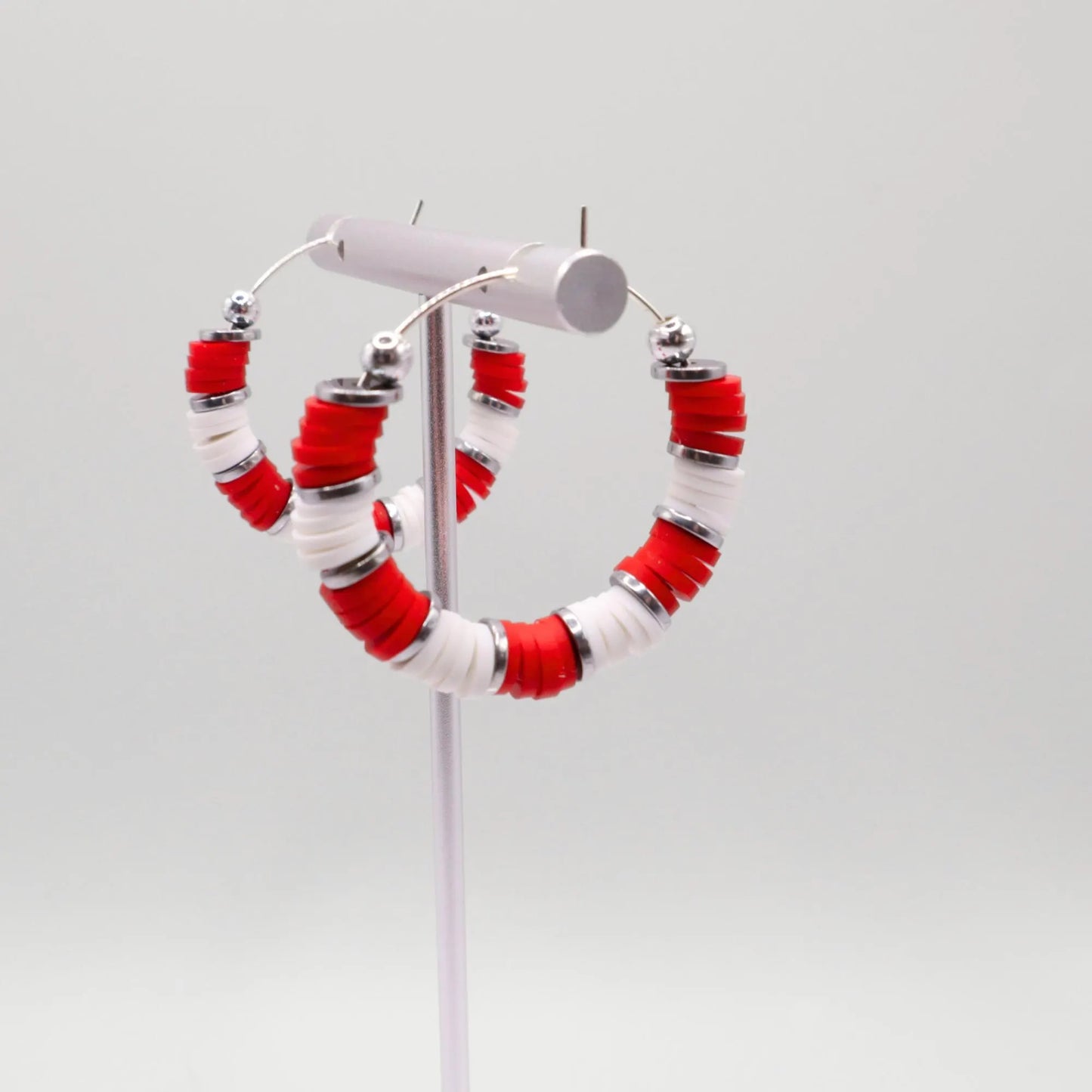 Side angle view of red and white heishi bead hoop earrings with silver spacers on a display stand against a white background