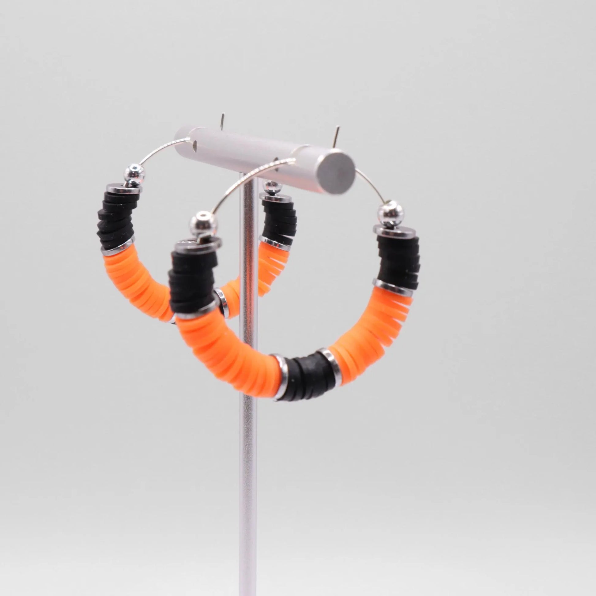 Orange and black heishi bead hoop earrings with silver accents hanging on a minimalist jewelry stand against a white background