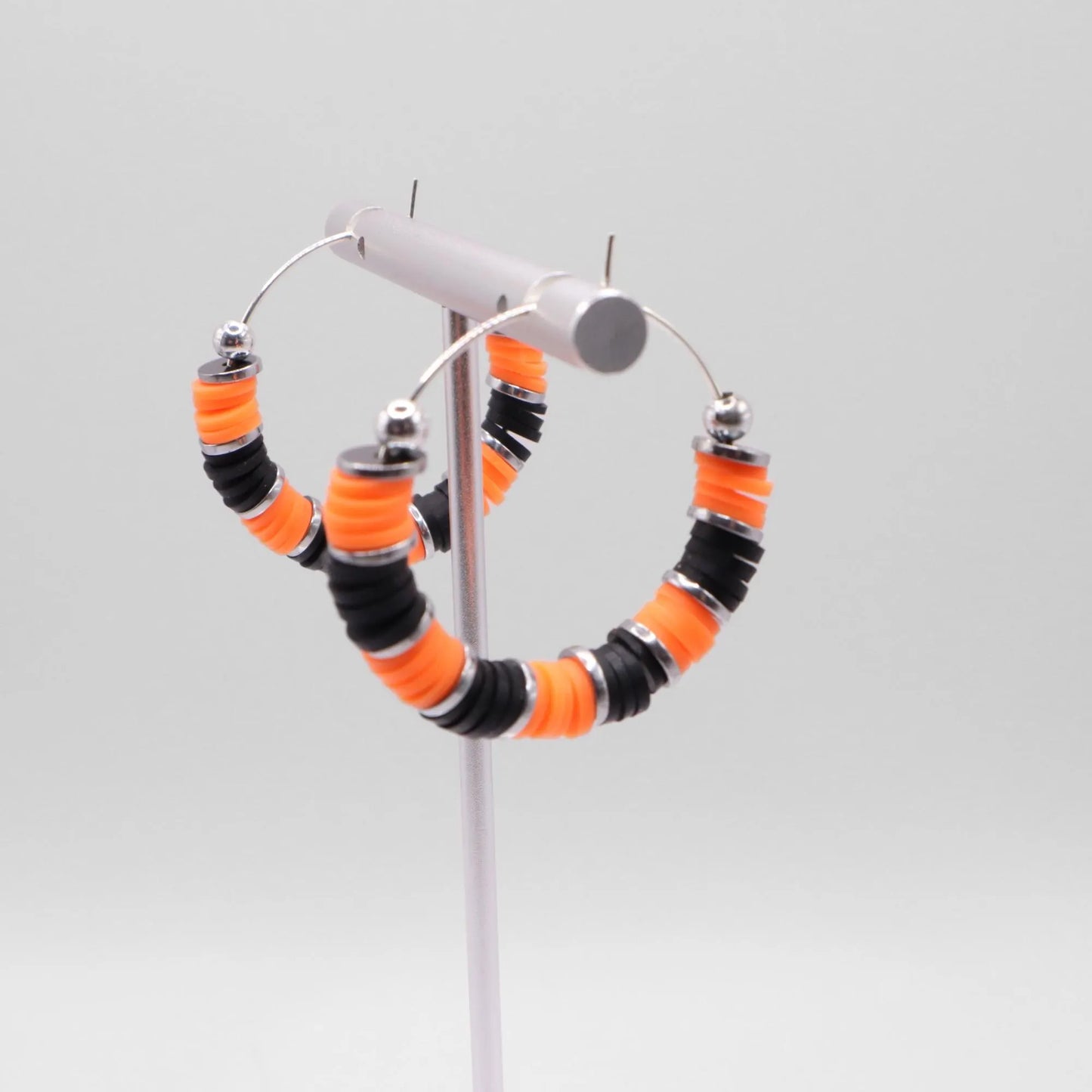 Close-up of orange and black heishi bead hoop earrings with silver accents on a jewelry stand, highlighting bead detail and pattern