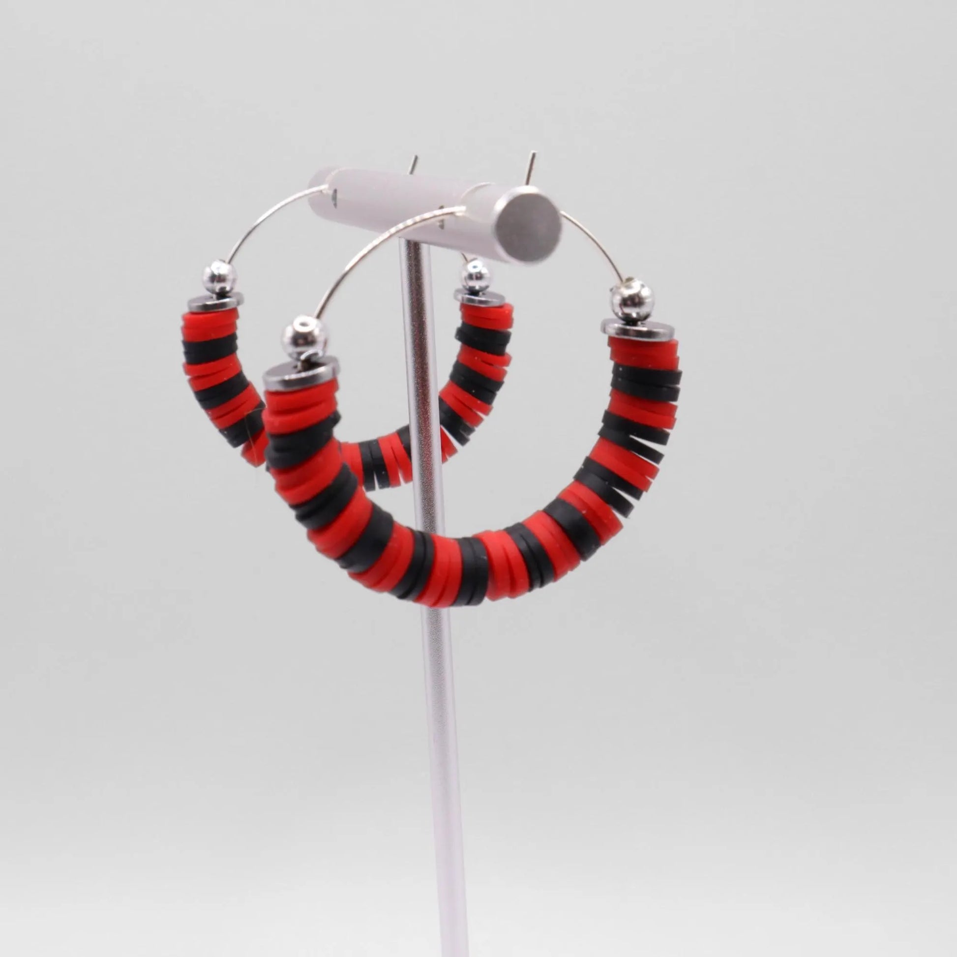 Red and black heishi bead hoop earrings with silver accents hanging on a minimalist jewelry stand against a white background