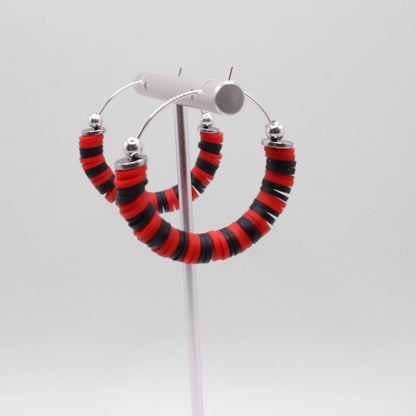 Red and black heishi bead hoop earrings with silver accents hanging on a minimalist jewelry stand against a white background