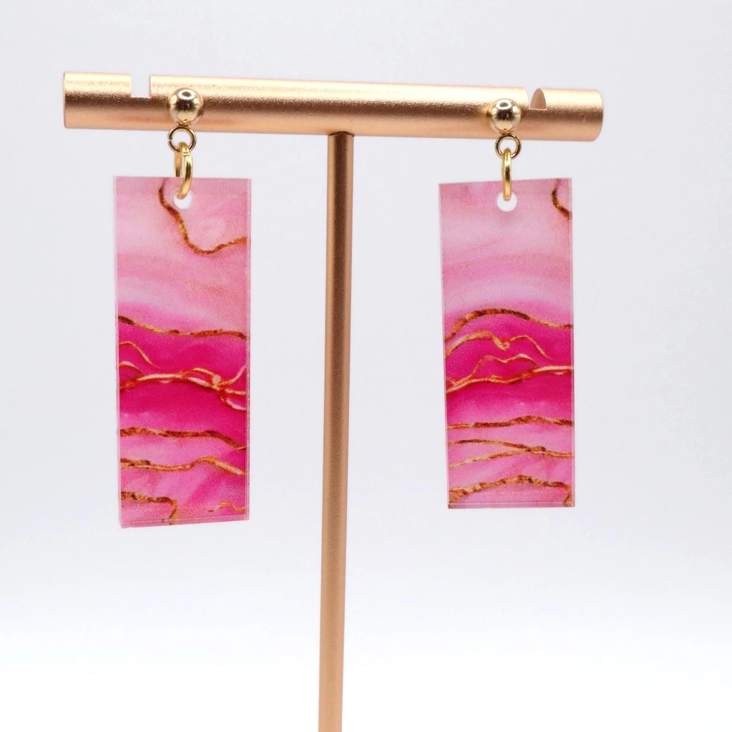 Pink marbled earrings with gold accents on a gold earring stand against a white background
