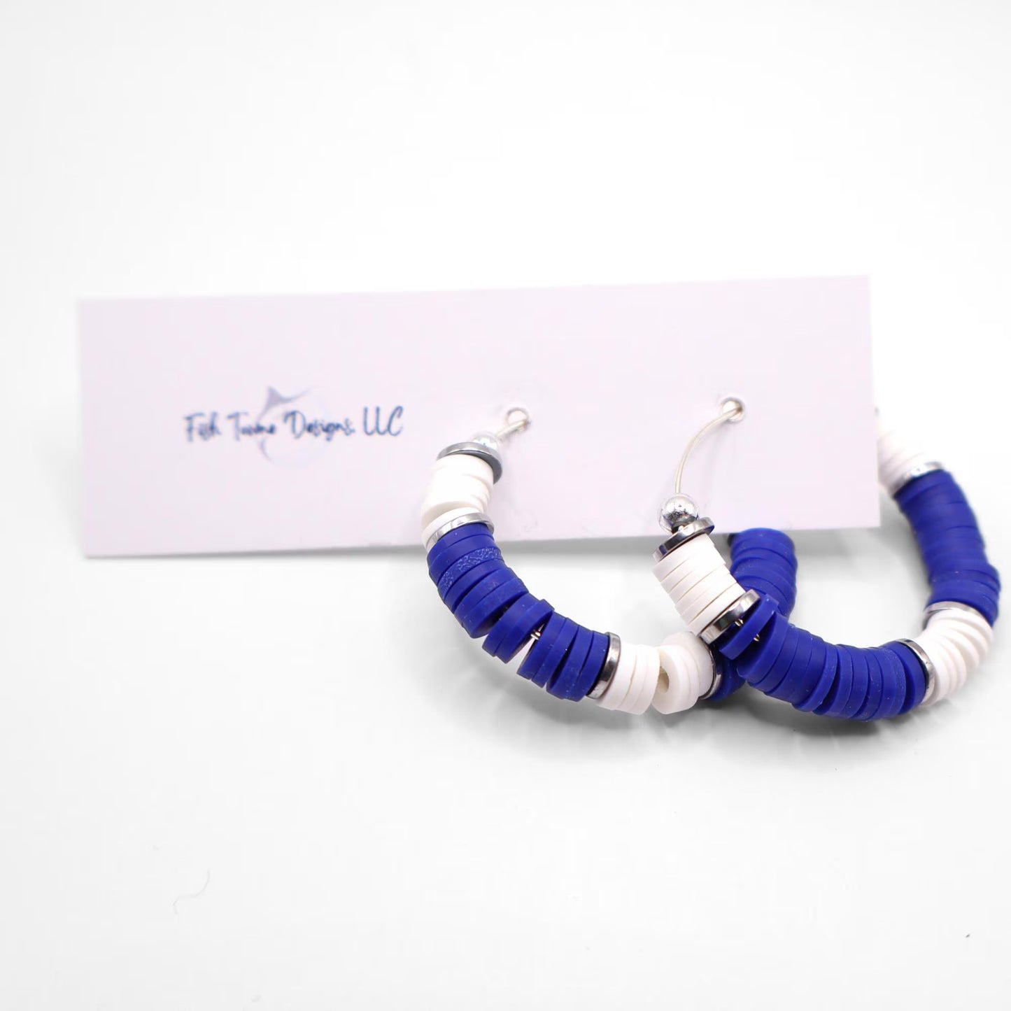 Royal blue and white heishi bead hoop earrings with silver accents hanging on a minimalist jewelry stand against a white background