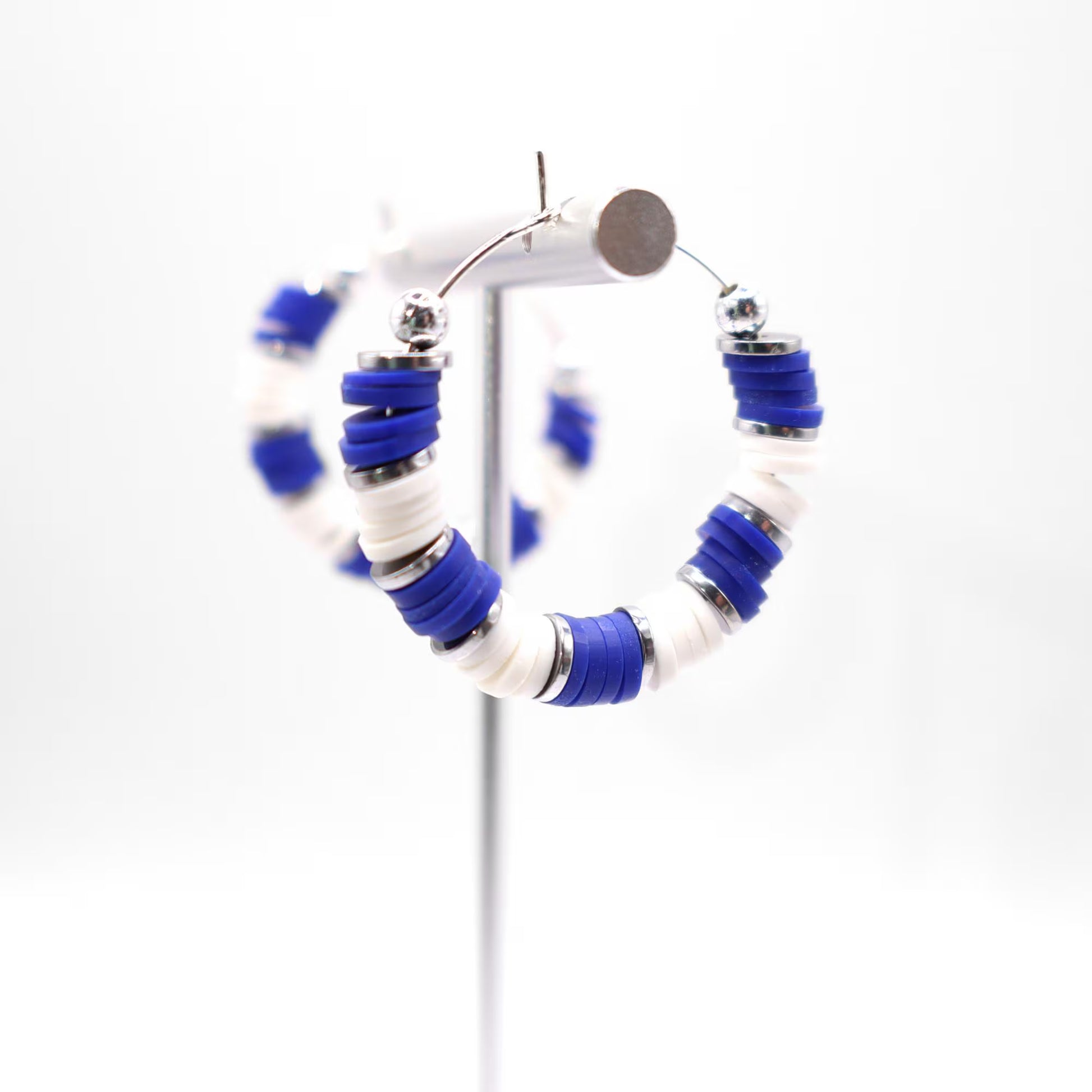 Side angle view of royal blue and white heishi bead hoop earrings with silver spacers on a display stand against a white background