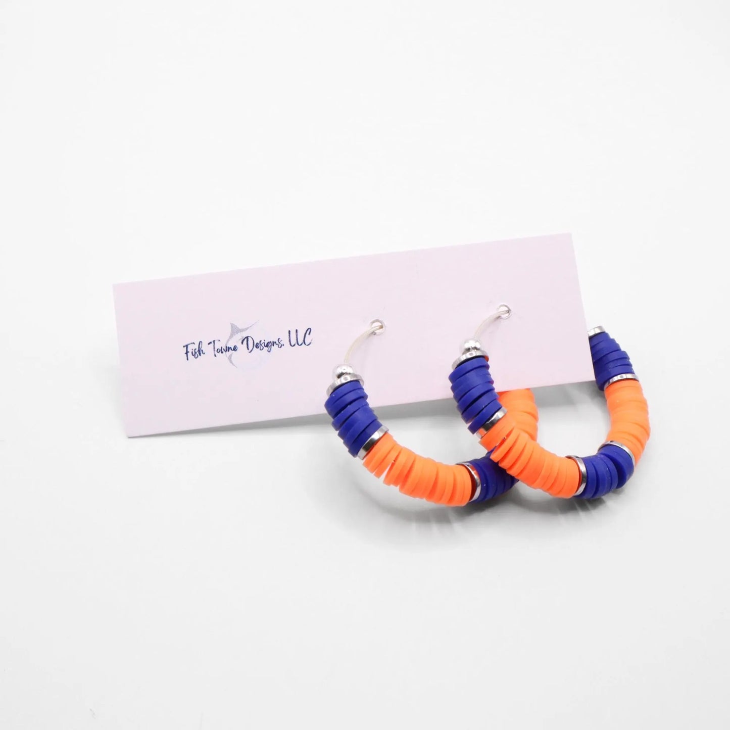 Blue and orange heishi bead hoop earrings with silver accents hanging on a minimalist jewelry stand against a white background