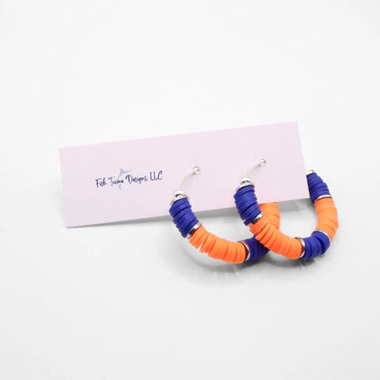 Blue and orange heishi bead hoop earrings with silver accents hanging on a minimalist jewelry stand against a white background