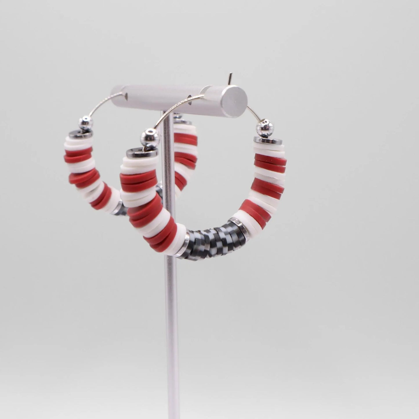Close-up of crimson and houndstooth heishi bead hoop earrings with silver accents on a jewelry stand, highlighting bead detail and pattern