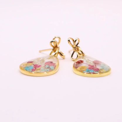 A pair of gold-colored earrings with a teardrop-shaped design. Each earring features a clear resin filled with colorful dried flowers and an 18k gold plated bow-shaped top. The earrings are set against a white background.