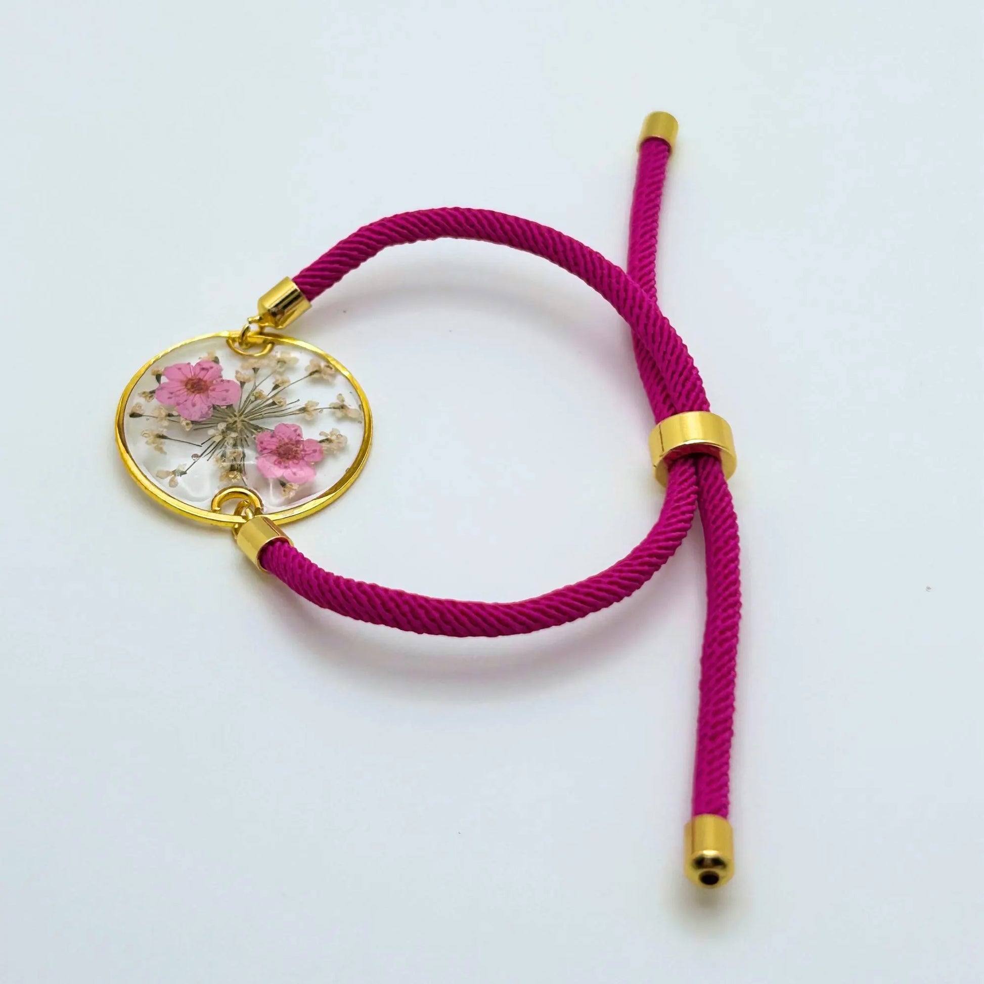 A magenta adjustable bracelet with a gold-colored circular pendant. The pendant contains pink flowers and white accents encased in clear resin. The bracelet's cord and metal accents are gold-colored.