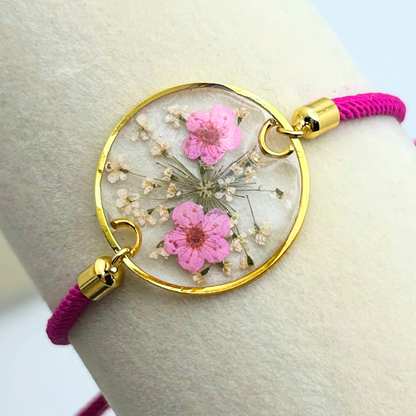 A hot pink adjustable bracelet with a gold-colored circular pendant. The pendant is filled with dried pink flowers and clear resin. The bracelet's cord is a vibrant pink, and the adjustable clasp is gold-colored.